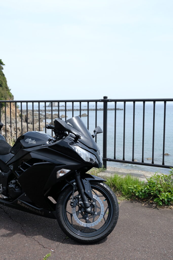 海辺の道路脇に停車した黒いKawasaki NINJA250、背景に岩場と青い海が広がる